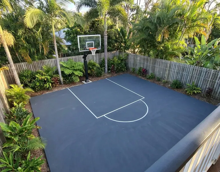 Backyard basketball half-court in dark blue with white lines and a hoop, surrounded by tropical plants